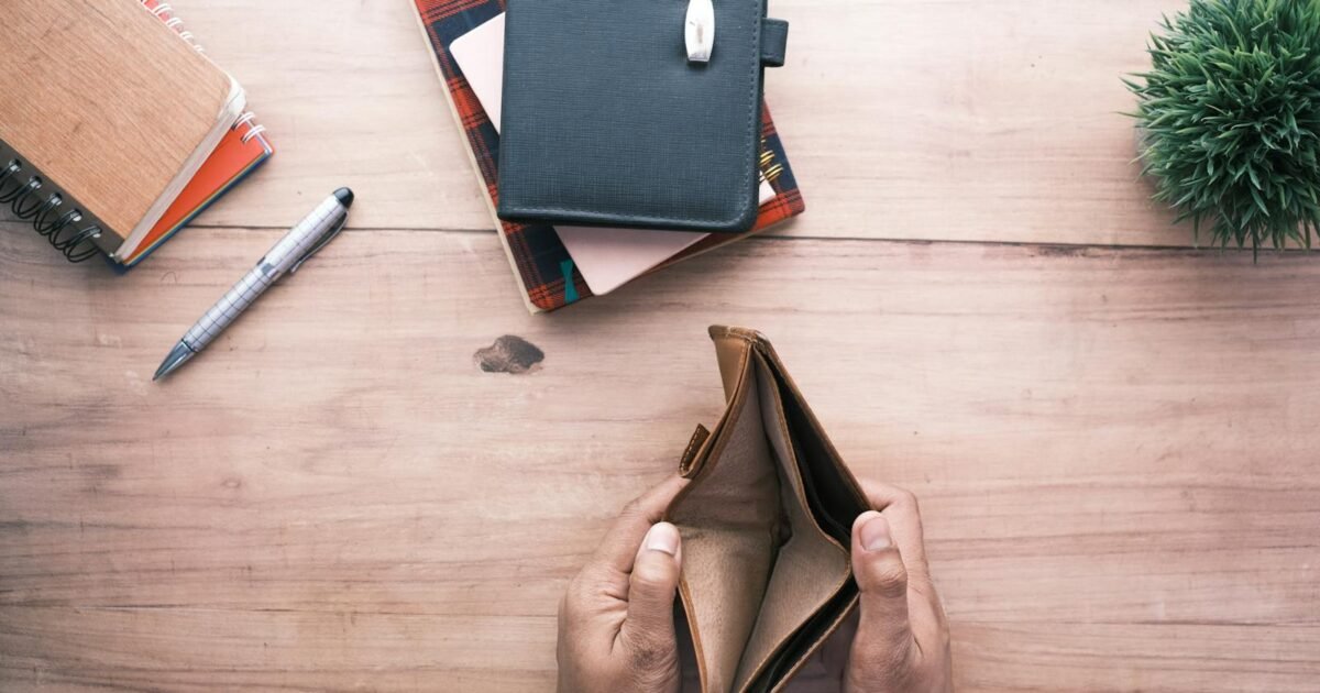 Top view of hands holding an open empty wallet on a wooden desk with notebooks and pen.