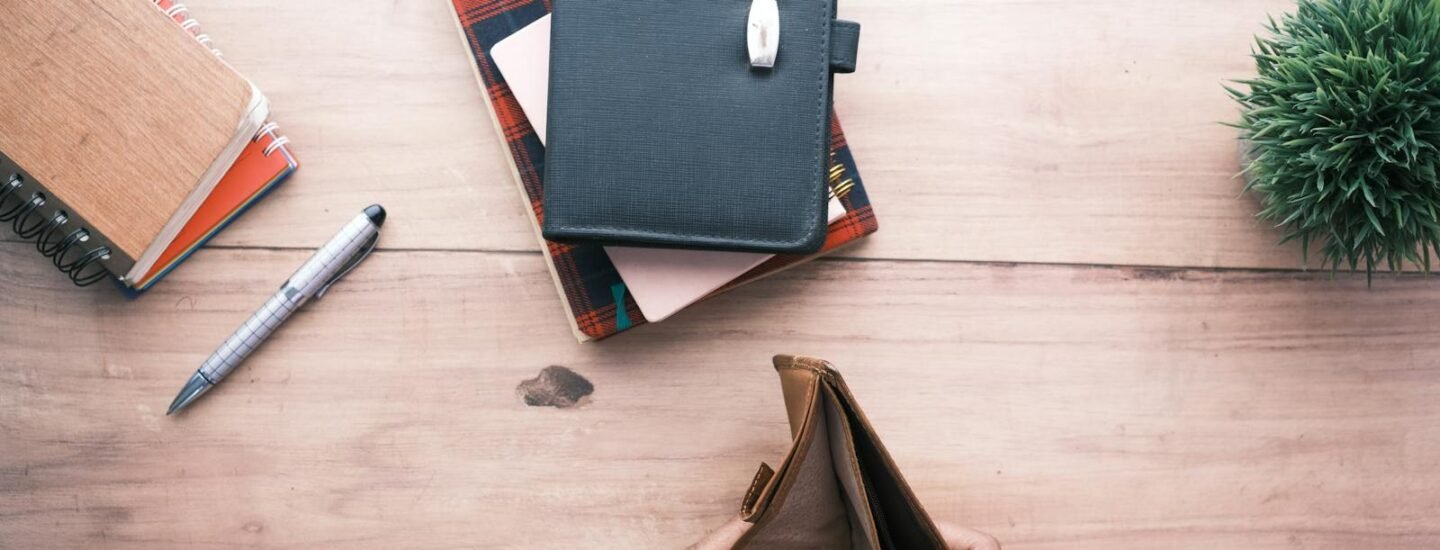 Top view of hands holding an open empty wallet on a wooden desk with notebooks and pen.