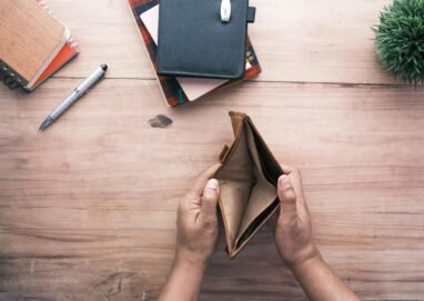 Top view of hands holding an open empty wallet on a wooden desk with notebooks and pen.