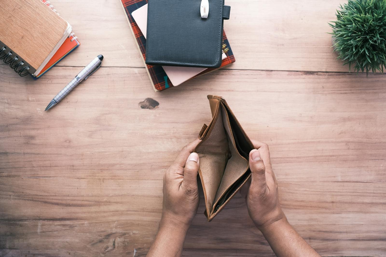 Top view of hands holding an open empty wallet on a wooden desk with notebooks and pen.