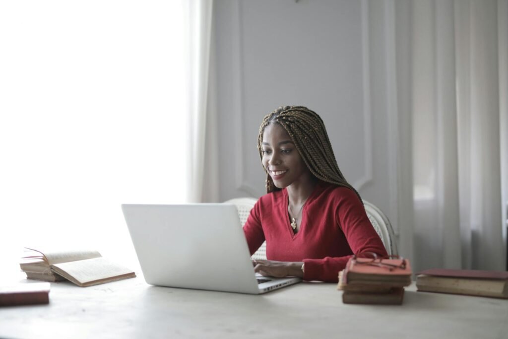 Woman with braided hair smiling as she works on a laptop at home with books around her.