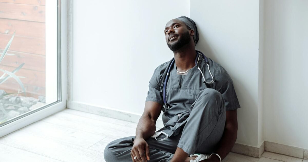 A tired healthcare professional in gray scrubs resting against a wall, reflecting exhaustion.