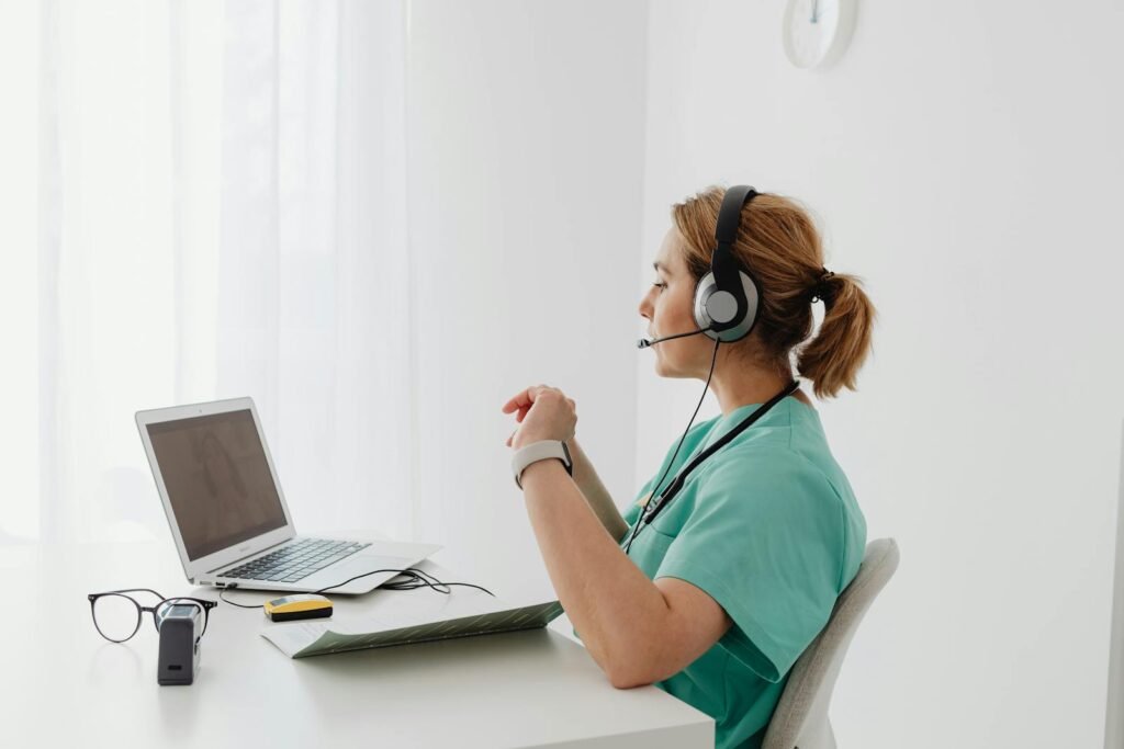 A female doctor using a laptop for an online consultation, wearing a headset in a bright office.