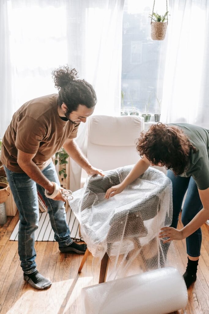 A couple is securing furniture with bubble wrap while packing in a bright, sunlit room.