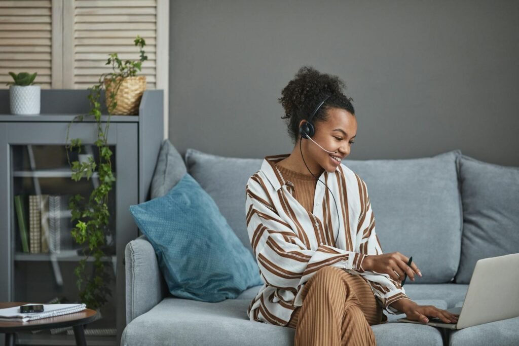 African American woman with a headset working from home on a laptop, sitting on a sofa.