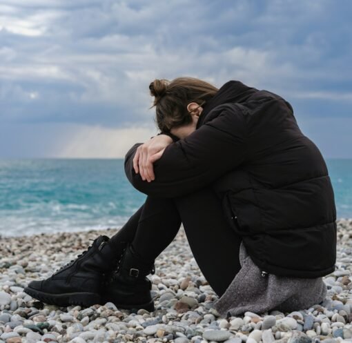 a woman sitting on a rocky beach with her head in her hands